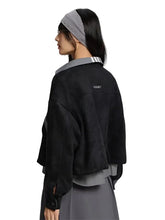 A model shown from the back wearing a black cropped suede-like jacket over a gray collared shirt and a gray skirt, with a gray beaded headband. The jacket features a small rectangular brand patch on the upper back.