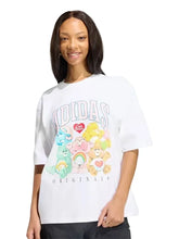 A woman wearing a white Adidas Originals graphic t-shirt featuring Care Bears characters, set against a solid light gray background.