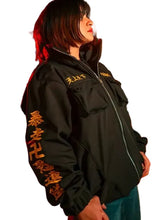 A person standing outdoors at night wearing a black zip-up jacket with large gold Chinese characters printed down one sleeve and across the chest, paired with blue jeans.