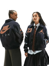 Two models wearing matching dark navy varsity-style jackets with orange Nike and Jordan logos, shown against a light gray studio background.