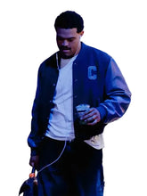A man wearing a navy blue varsity jacket with a light blue 'C' on the chest, a white t-shirt, and dark pants, holding a drink against a stylized, graffiti-textured wall.