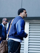 A man wearing a blue leather-look bomber jacket, dark cargo pants, and a brown shoulder bag walks outdoors next to a gray building with louvered windows. Another man in sunglasses is partially visible in the background.