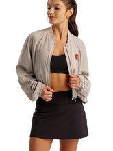 A woman modeling a light beige athletic bomber jacket with a small embroidered logo on the left chest, worn over a black sports bra and black tennis skirt against a solid gray background.