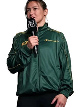 A woman wearing a dark green Champion track jacket with gold piping and logo, holding a black microphone with a DAZN logo, against a solid light gray background.