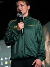 A woman on stage wearing a dark green Champion track jacket with gold piping and logo, holding a black microphone with a DAZN logo.