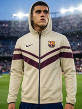 A man wearing a cream-colored FC Barcelona track jacket with dark purple stripes and a navy blue Nike swoosh, standing on a soccer field with stadium lights in the background.