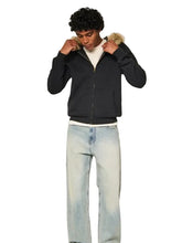A man wearing a black zip-up hoodie with a beige faux-fur trimmed hood, light-wash blue jeans, and a white t-shirt, standing against a plain off-white studio background.