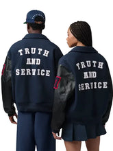 Two models shown from the back wearing matching navy blue varsity-style jackets with black leather sleeves and large white text reading 'TRUTH AND SERVICE' across the back. One model also wears a navy cap, and both are photographed against a plain light gray studio background.