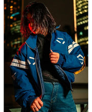 A person wearing a blue bomber jacket with white graphic stripes and a black shirt, posed against a blurred nighttime cityscape background.