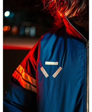 A close-up of a person wearing a blue zip-up jacket with reflective silver triangular patches on the chest and reflective orange-and-white stripes on the sleeve, photographed at night with dramatic red and blue lighting.