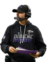 A man wearing a black Baltimore Ravens hoodie with 'DAIENCE ENGINEERED FOR' text, a black cap, and a Sony headset, holding purple folders against a light gray background.