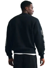A man wearing a black crewneck sweatshirt with white graphic prints on the sleeves, shown from the back against a plain light background.
