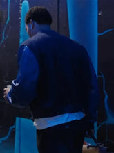 A man viewed from behind wearing a dark blue and black varsity-style jacket over a white shirt, standing in a dimly lit environment with blue lighting.