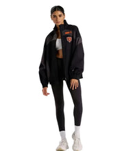 A woman modeling a black athletic jacket with gray side panels and orange logo details, paired with black leggings, white socks, and white sneakers, against a solid light gray background.