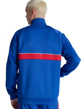 A man viewed from the back wearing a royal blue track jacket with a bold red horizontal stripe across the upper back, paired with matching blue track pants against a light gray studio background.