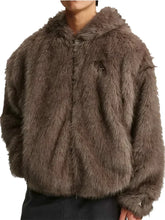 A person wearing a shaggy faux-fur cropped jacket in a taupe-brown color, paired with dark pants, against a light gray studio background.