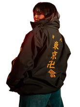 A person standing on a city street at night wearing an oversized black jacket with large gold Japanese characters printed down the back, paired with blue jeans.