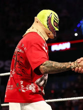 A wrestler in a bright neon yellow mask with black and red details, a red graphic t-shirt, and white pants shakes hands inside a wrestling ring under arena lights.