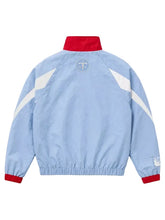 A light blue windbreaker jacket shown from the back, featuring a red collar and cuffs, white diagonal shoulder stripes, and a circular 'TT' logo on the upper back.