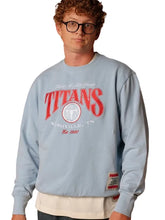 A man wearing a light blue sweatshirt with a red and white 'Titans' graphic on the front, paired with a cream undershirt and black glasses, against a light gray background.
