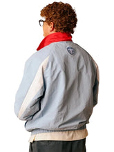 A person shown from the back wearing a light blue color-blocked jacket with cream and red panels, a red neck collar, and a circular logo patch on the upper back.