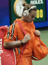 A woman wearing an orange tracksuit and white headphones walks on a tennis court, carrying an orange tennis bag and a red tennis racket. A Rolex sign is visible in the background.
