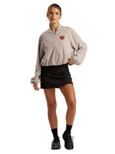 A woman modeling a light beige zip-up windbreaker jacket with a small red and black shield logo on the left chest, paired with a black athletic mini skirt, black socks, and black sneakers, against a solid light gray studio background.